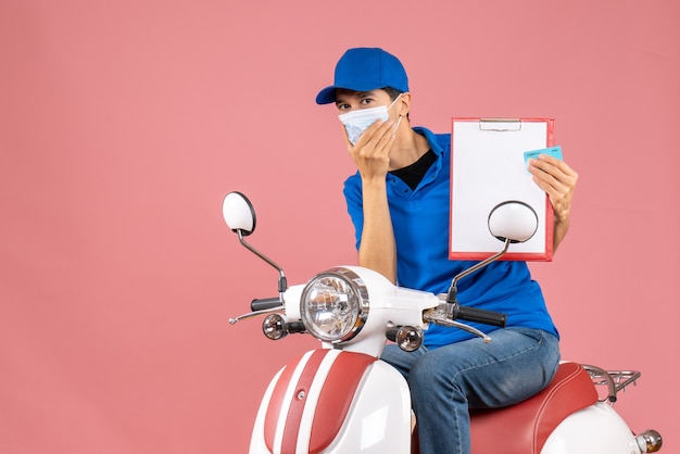 Draufsicht auf einen überraschten männlichen Lieferer in Maske mit Hut, der auf einem Roller sitzt und ein Dokument und eine Bankkarte auf pastellfarbenem Pfirsich zeigt