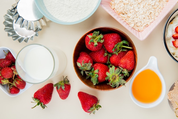 Draufsicht auf Erdbeeren in Schüssel und Glas Milch und geschmolzene Butter mit Mehl und Hafer auf Weiß