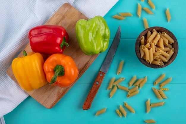 Draufsicht auf farbige Paprika auf einem Schneidebrett mit rohen Nudeln und einem Messer auf einer türkisfarbenen Oberfläche