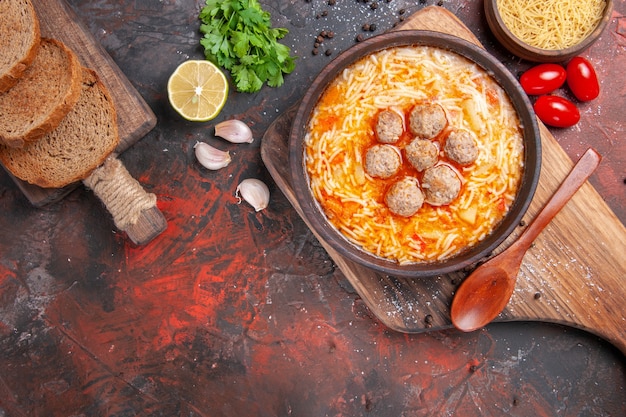 Draufsicht auf Fleischbällchensuppe mit Nudeln ungekochte Pasta Schneidebrett Zitrone ein Bündel grüne Tomaten verschiedene Gewürze auf dunklem Tisch