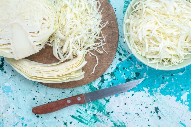 Draufsicht auf frisch geschnittenen Kohl mit halb ganzem Gemüse mit Messer auf hellblauem Schreibtisch, Gemüsesalatmahlzeit-gesunder Salat