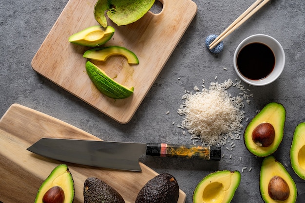 Draufsicht auf frische Avocados, ein Schneidebrett und ein Messer, Reis und Stäbchen auf einer grauen Oberfläche