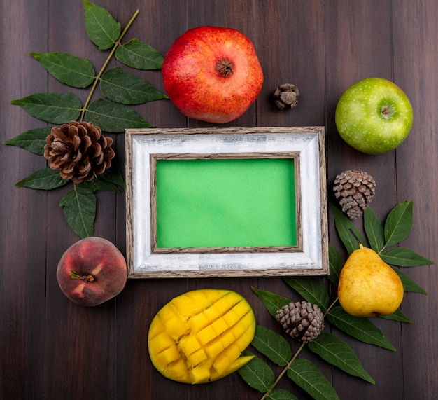 Draufsicht auf frische Früchte wie Granatapfel-grüne Apfelbirnen-geschnittene Mango, isoliert mit Blatt- und Tannenzapfen auf Holz