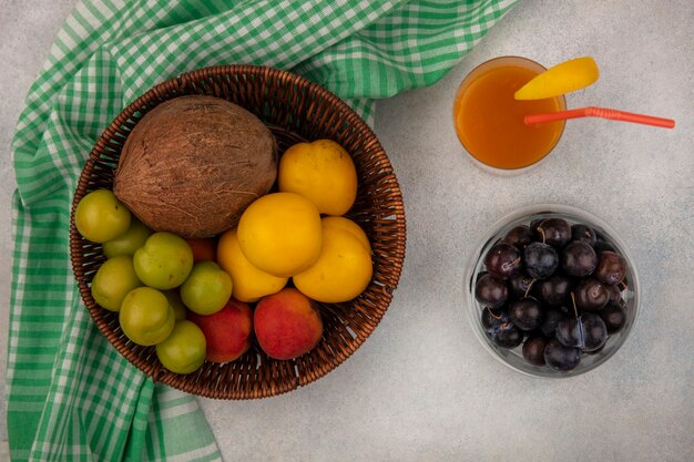 Draufsicht auf frische Früchte wie Kokosnusspfirsichgrüne Kirschpflaumen auf einem Eimer auf einem karierten Tuch mit Schlehen auf einer Schüssel mit frischem Pfirsichsaft auf einem weißen Hintergrund