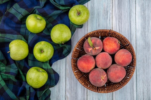 Draufsicht auf frische Früchte wie Äpfel auf karierter Tischdecke und Pfirsiche auf Eimer auf grauem Holz