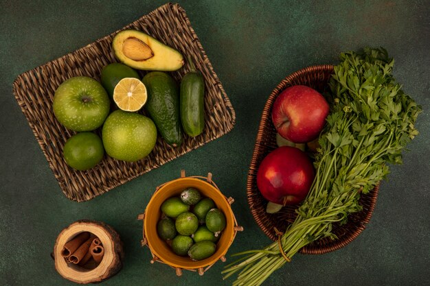 Draufsicht auf frische Lebensmittel wie grüne Äpfel Avocados Gurke auf einem Weidentablett mit Feijoas auf einem Eimer mit roten Äpfeln und Petersilie auf einem Eimer auf einer grünen Wand