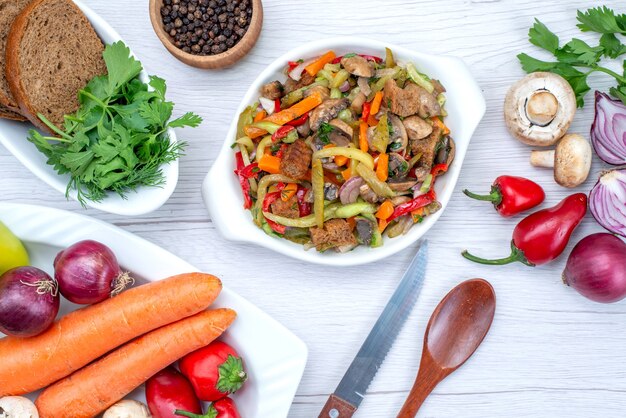 Draufsicht auf frischen Gemüsesalat, der mit Fleisch zusammen mit Brotlaib und ganzem Gemüse und Gemüse auf hellem Schreibtisch geschnitten wird