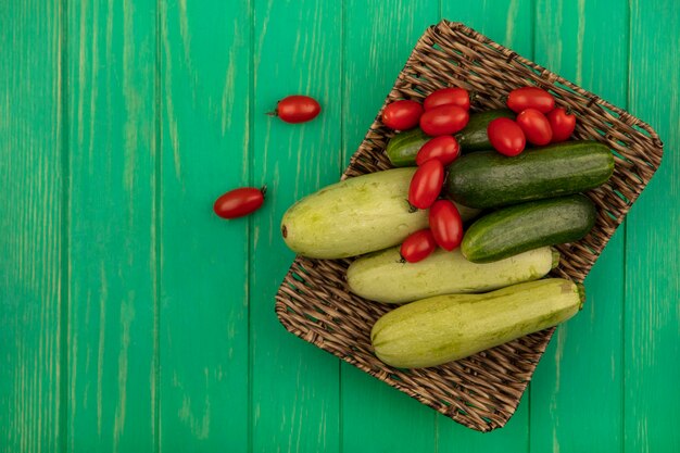 Draufsicht auf frisches Gemüse wie Pflaumentomatengurken und Zucchini auf einem Weidentablett auf einer grünen Holzwand mit Kopierraum