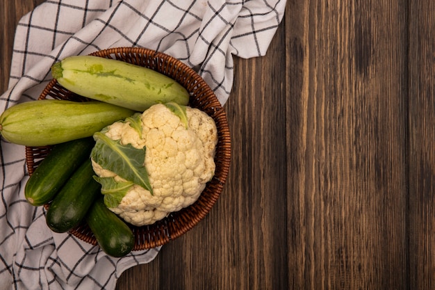 Draufsicht auf frisches Gemüse wie Zucchini-Gurken-Blumenkohl auf einem Eimer auf einem karierten Tuch auf einer Holzwand mit Kopierraum