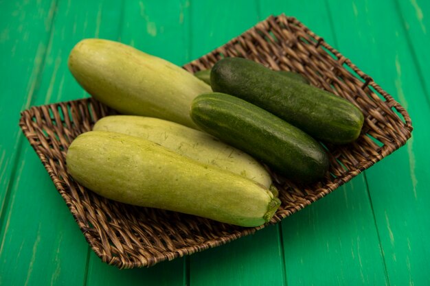 Draufsicht auf frisches und gesundes Gemüse wie Gurken und Zucchini auf einem Weidentablett auf einer grünen Holzwand