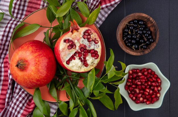 Draufsicht auf Früchte als Granatapfelhälfte halb und ganz mit Blättern auf kariertem Stoff und Schalen mit Schlehen- und Granatapfelbeeren auf schwarzer Oberfläche