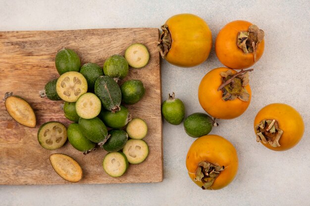 Draufsicht auf ganze und halbierte frische Feijoas lokalisiert auf einem hölzernen Küchenbrett mit Kakis auf einer grauen Wand