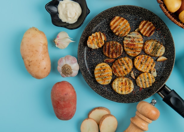 Draufsicht auf gebratene Kartoffelscheiben in der Pfanne mit ungekochtem Mayonnaise-Knoblauchsalz auf Blau