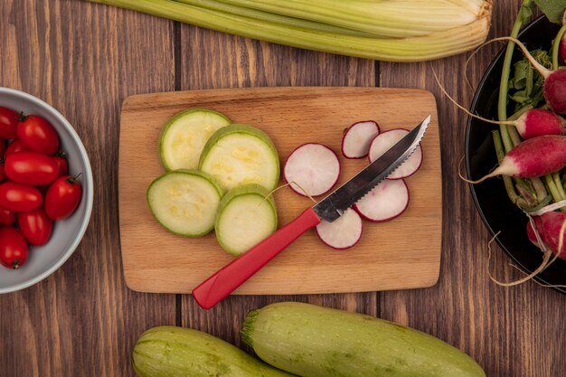 Draufsicht auf gehackte Scheiben Zucchini und Radieschen auf einem hölzernen Küchenbrett mit Messer mit Pflaumentomaten auf einer Schüssel mit Radieschen auf einer Schüssel mit Zucchini und Sellerie lokalisiert auf einem hölzernen Hintergrund