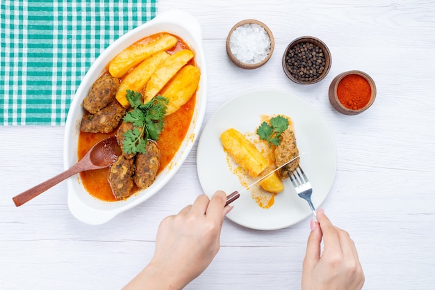 Draufsicht auf gekochte Fleischkoteletts mit Soßenkartoffeln und grünem Essen durch Frau auf leichtem Schreibtisch, Lebensmittelmahlzeit-Fleischgemüse