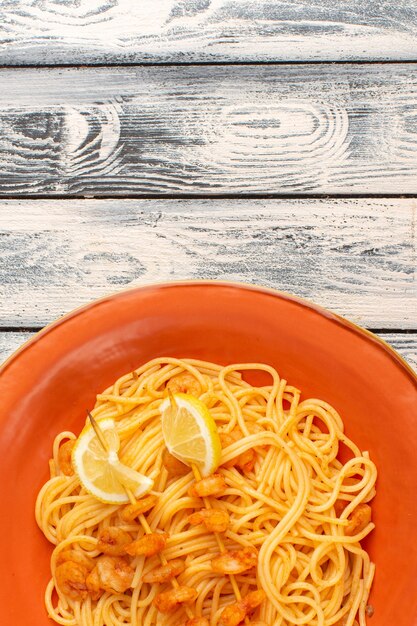 Draufsicht auf gekochte italienische Nudeln, die mit Zitronenscheiben und Garnelen innerhalb des orangefarbenen Tellers auf der grauen rustikalen Holzoberfläche lecker sind