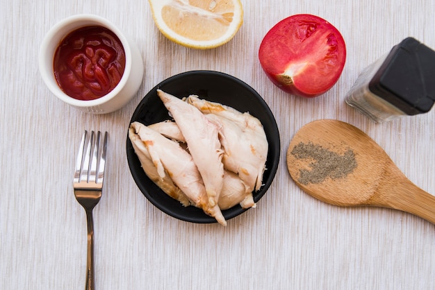 Draufsicht auf gekochtes Hähnchen in Schwarzblech mit Pulver aus schwarzem Pfeffer; Tomate; Zitrone; Gabel und Tomatensauce über Schreibtisch aus Holz