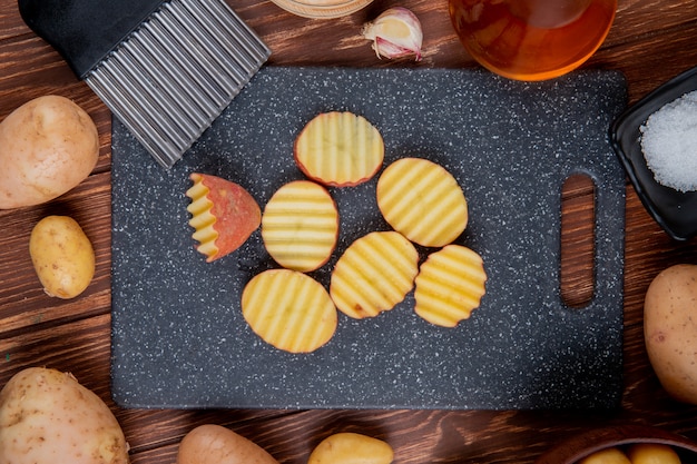 Kostenloses Foto draufsicht auf gekräuselte kartoffelscheiben auf schneidebrett mit ganzen knoblauchbutter und salz auf holz herum