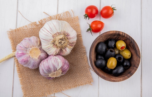 Draufsicht auf Gemüse als Knoblauchknollen auf Sackleinen und Olivenschüssel mit Tomaten auf Holzoberfläche