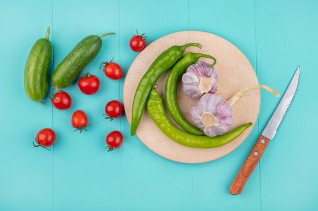 Draufsicht auf Gemüse als Pfeffer und Knoblauch auf Schneidebrett mit Gurkentomate und Messer auf blauer Oberfläche