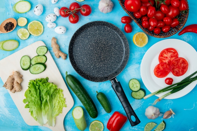 Draufsicht auf Gemüse wie Salat, Gurke, Ingwer und andere mit Pfanne und Zitrone auf blauer Oberfläche