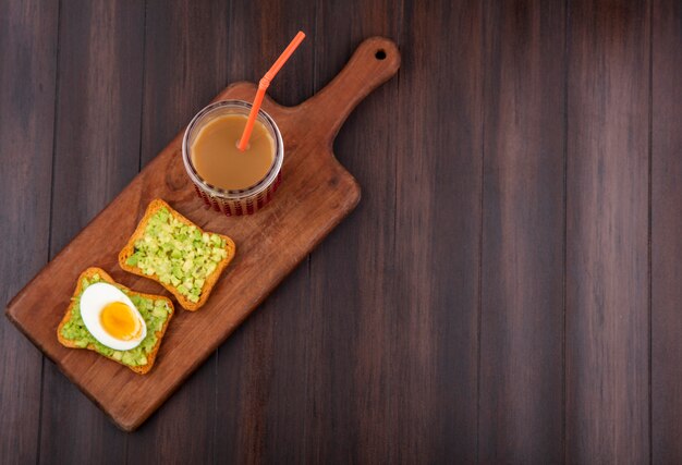 Draufsicht auf geröstete Brotscheiben mit Avocadopulpen und Ei mit Saft in einem Glas auf einem hölzernen Küchenbrett auf einer hölzernen Oberfläche