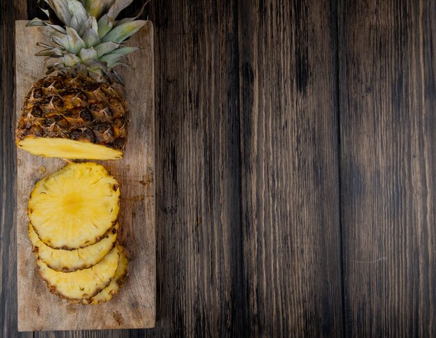 Draufsicht auf geschnittene und geschnittene Ananas auf Schneidebrett auf der linken Seite und hölzernem Hintergrund mit Kopienraum