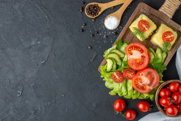 Draufsicht auf geschnittenen ganzen frischen Tomaten- und Gurkenkäse auf Holzbrettbesteck stellte Gewürze in Löffel auf schwarzer Oberfläche ein