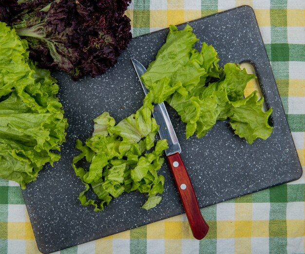 Draufsicht auf geschnittenen Salat mit Messer auf Schneidebrett und ganze mit Basilikum auf kariertem Stoff