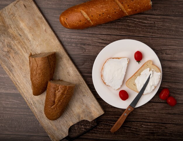 Draufsicht auf geschnittenes halbes Baguette und Weißbrotscheiben mit Tomaten und Messer in Platte auf hölzernem Hintergrund