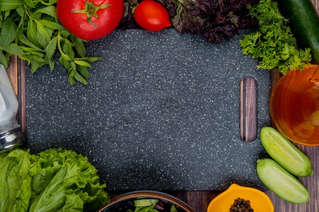 Draufsicht auf geschnittenes und ganzes Gemüse als Tomaten-Basilikum-Minze-Gurken-Salat-Koriander mit schwarzem Salzpfeffer und Schneidebrett auf Holzoberfläche