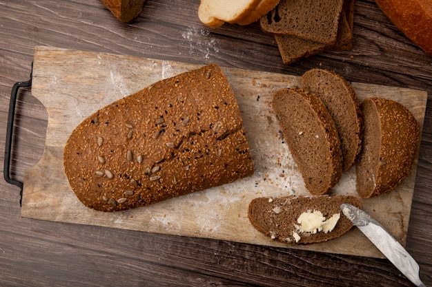 Draufsicht auf geschnittenes und geschnittenes Sandwichbrot und Brotscheibe mit Butter darauf und Messer auf Schneidebrett auf hölzernem Hintergrund