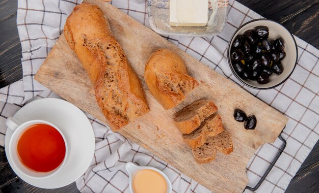 Draufsicht auf geschnittenes und geschnittenes schwarzes Baguette mit einer Tasse Teebutterolive auf kariertem Stoff und Holztisch