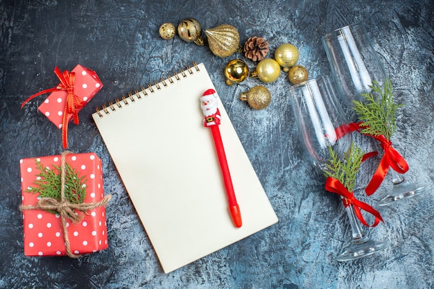 Draufsicht auf Glasbecher mit rotem Band und Dekorationszubehör neben Geschenkboxen-Notizbuch mit Stift auf dunklem Hintergrund