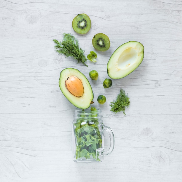 Draufsicht auf halbierte Avocado; Kiwi; Rosenkohl und Salat mit Glas vor hölzernen Hintergrund