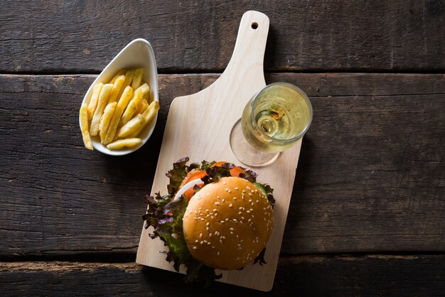 Draufsicht auf hausgemachte Hähnchenburger mit Champagner, Pommes frites, Salat, Tomaten und Zwiebeln auf Holzbrett.