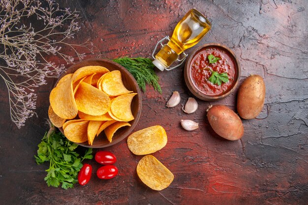 Draufsicht auf hausgemachte köstliche knusprige Kartoffelchips in einer kleinen braunen Schüssel Kartoffeln Ölflasche grüne Tomaten Knoblauch und Ketchup auf dunklem Hintergrund