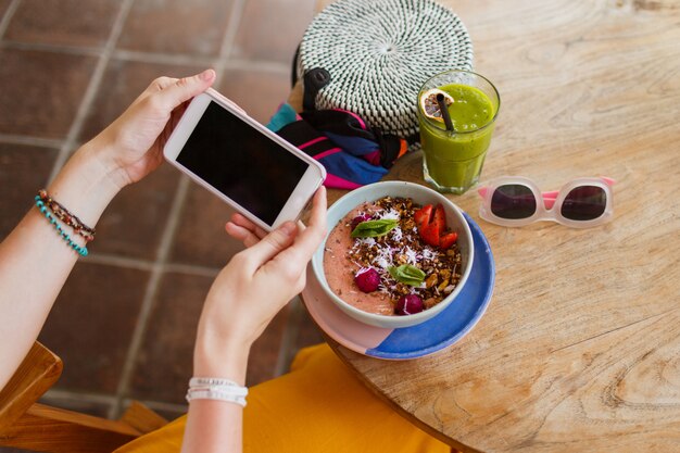 Draufsicht auf hübsche Frau in der gelben Hose, die Telefon benutzt und leckeres veganes Essen genießt