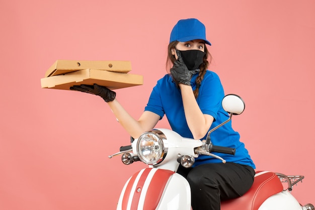 Draufsicht auf junge fokussierte Kurierinnen mit medizinischer Maske und Handschuhen, die auf einem Roller sitzen und Bestellungen auf Pastellpfirsich liefern