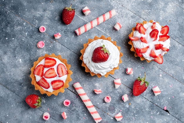 Draufsicht auf kleine cremige Kuchen mit geschnittenen und frischen Erdbeeren zusammen mit Stockbonbons auf grauem Schreibtisch