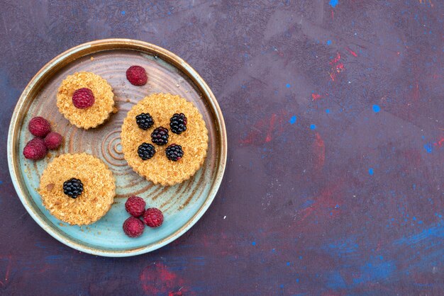 Draufsicht auf kleine Kuchen, die süß und köstlich mit Beeren auf der dunklen Oberfläche sind