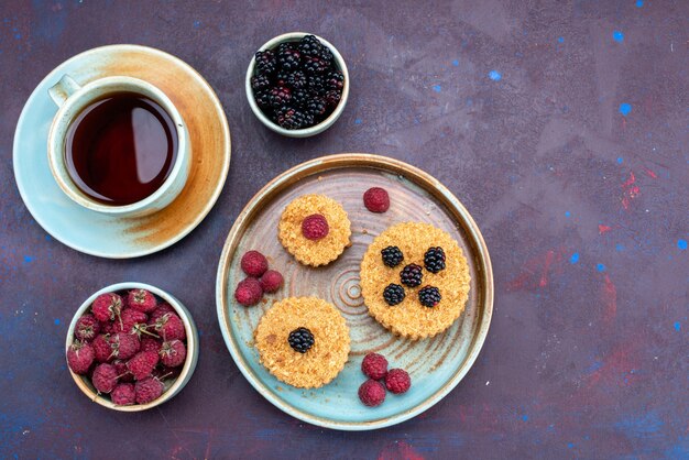 Draufsicht auf kleine Kuchen, die süß und köstlich mit frischen Beeren und Tee auf der dunklen Oberfläche sind