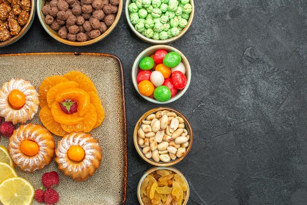 Draufsicht auf kleine leckere Kuchen mit Süßigkeiten, Früchten und Nüssen auf Grau