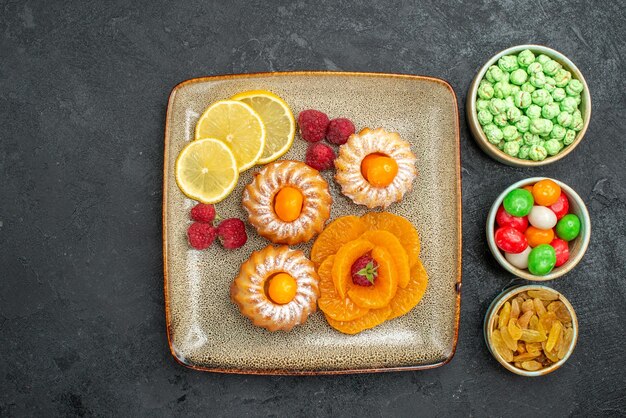 Draufsicht auf kleine leckere Kuchen mit Zitronenscheiben, Mandarinen und Bonbons auf Dunkelheit