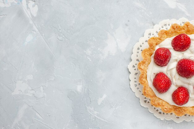 Draufsicht auf kleinen köstlichen Kuchen mit Sahne und frischen roten Erdbeeren auf hellem Schreibtisch