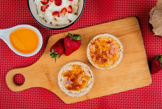 Draufsicht auf knusprige Knäckebrote, die mit Marmelade und Erdbeeren auf Schneidebrett mit Haferflocken und geschmolzener Butter auf Rot und Weiß verschmiert sind