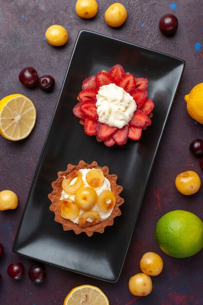 Draufsicht auf köstliche cremige Kuchen innerhalb Platte mit frischen Zitronen und Früchten auf der dunklen Oberfläche