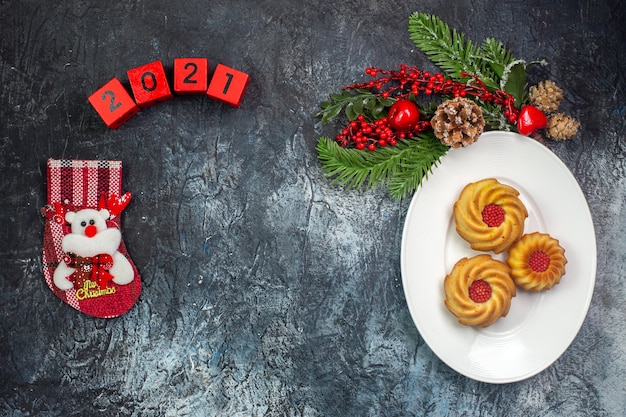 Draufsicht auf köstliche Kekse auf einem weißen Teller und Dekorationen Weihnachtsmann-Hutnummern Neujahrssocke auf dunkler Oberfläche