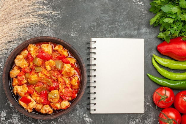 Draufsicht auf köstliche würzige Suppe mit Hühnchen und notwendigem Gemüse und einem Bündel Grün neben dem Notebook