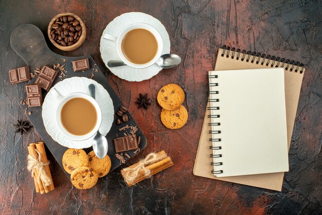 Draufsicht auf köstlichen Kaffee in weißen Tassen auf Holzschneidebrett Cookies Zimt Limetten Schokoriegel Spiralnotizbücher